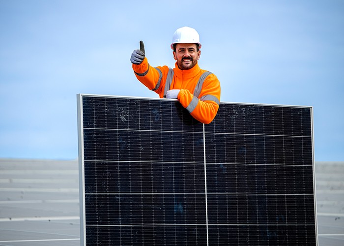 Worker in orange high-visibility jacket and hard hat giving a thumbs up behind a solar panel