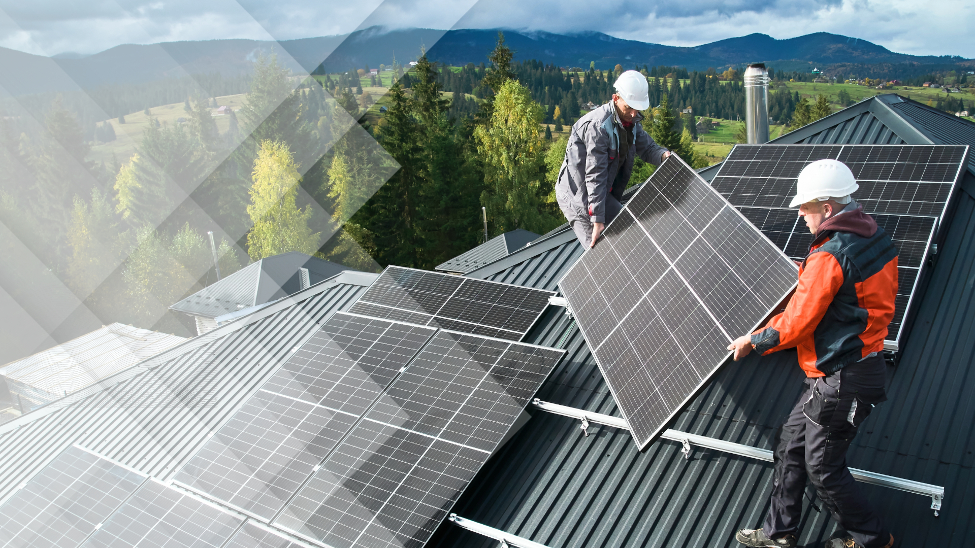 Two workers in hard hats carrying a solar panel across a metal roof with mountains behind
