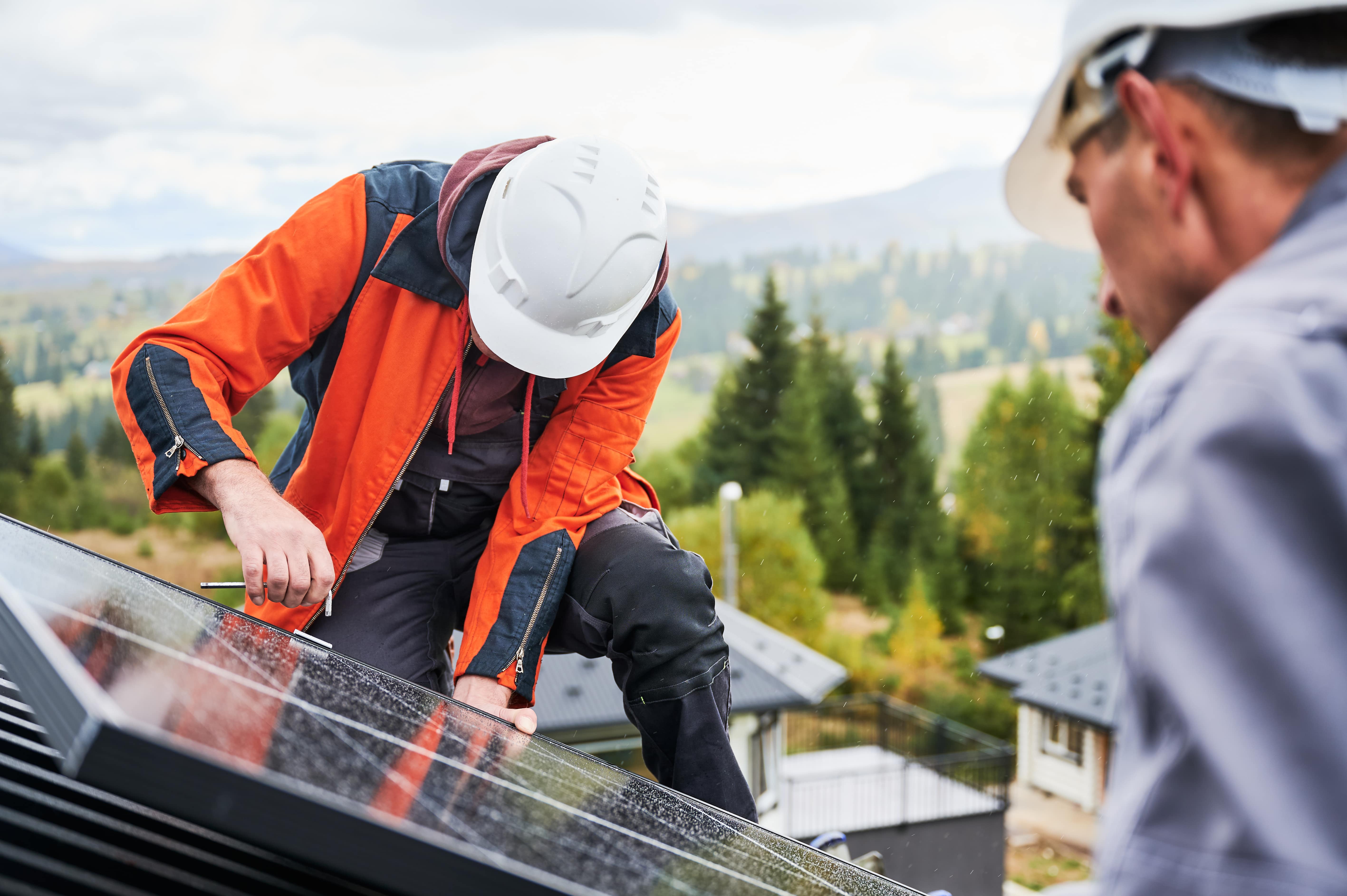 Installer in an orange jacket and white hard hat fastening a solar panel on a roof, second worker in foreground