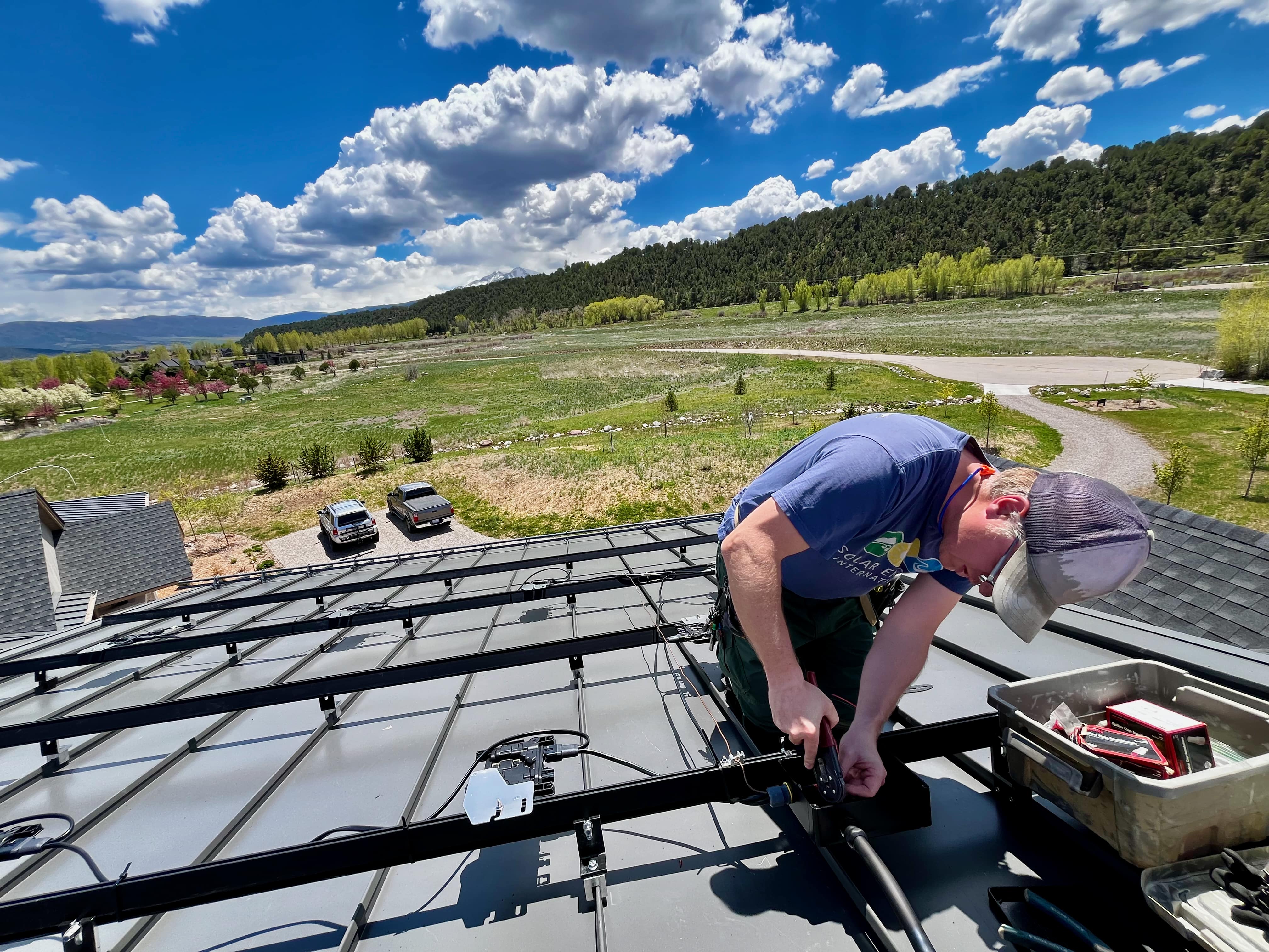 Installer kneeling on a metal roof wiring solar mounting rails above a green valley