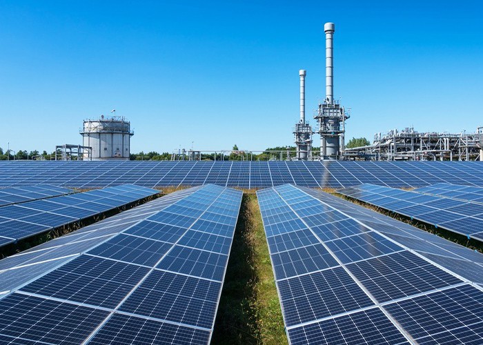 Rows of solar panels in a field with an industrial plant and tall stacks behind