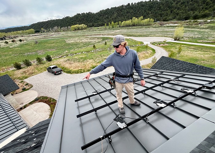 Installer standing on a standing-seam metal roof with solar mounting rails, a driveway and hills below