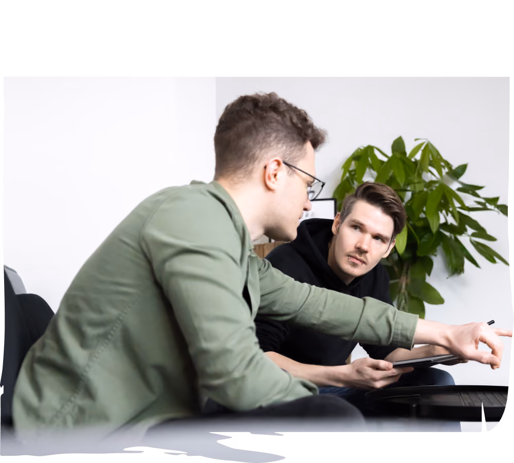 Zwei Männer sitzen in einem Büro und diskutieren etwas, während einer von ihnen ein Tablet in der Hand hält. Im Hintergrund steht eine grüne Pflanze.