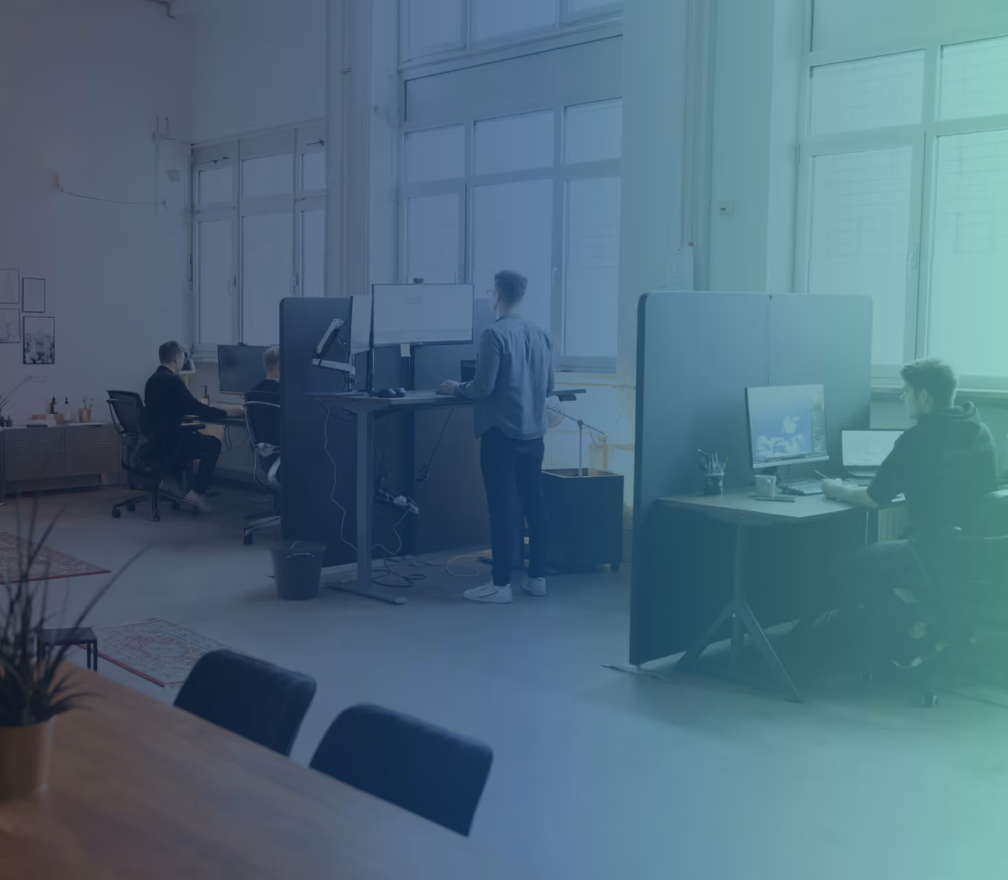 Büro mit mehreren Personen, einige sitzen an Schreibtischen, einer steht an Stehtisch mit Monitoren.