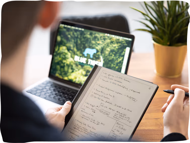 Person holding a stylus and writing notes on a tablet with a laptop showing a blue tiger logo in background.