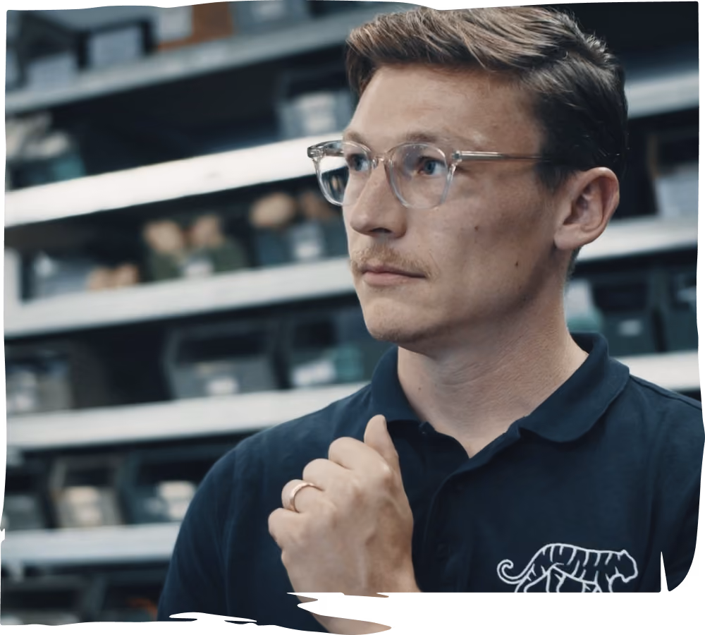 Ein Mann mit Brille und dunklem Polo mit Tigerlogo schaut nach rechts in einem Lagerraum.