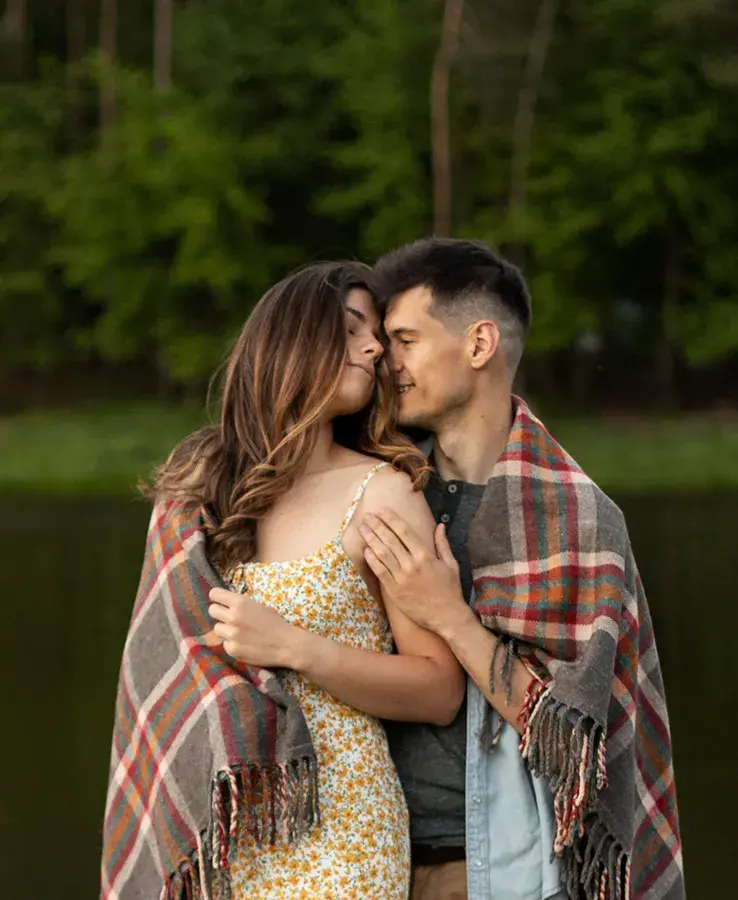 Una pareja joven y afectuosa de pie al aire libre, envuelta junta en una manta a cuadros. La mujer, con un vestido floral amarillo, tiene los ojos cerrados mientras el hombre se acurruca contra su mejilla. El fondo es un bosque verde junto a un lago.