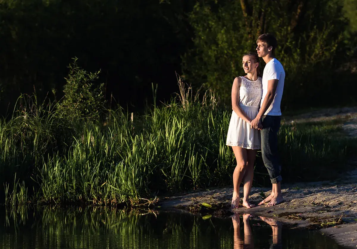 A young couple standing barefoot and holding hands on the sandy shore of a river or lake. They are illuminated by warm, late-afternoon light, looking off to the side. The background features tall grass and lush trees, and their reflection is visible in the calm water.
