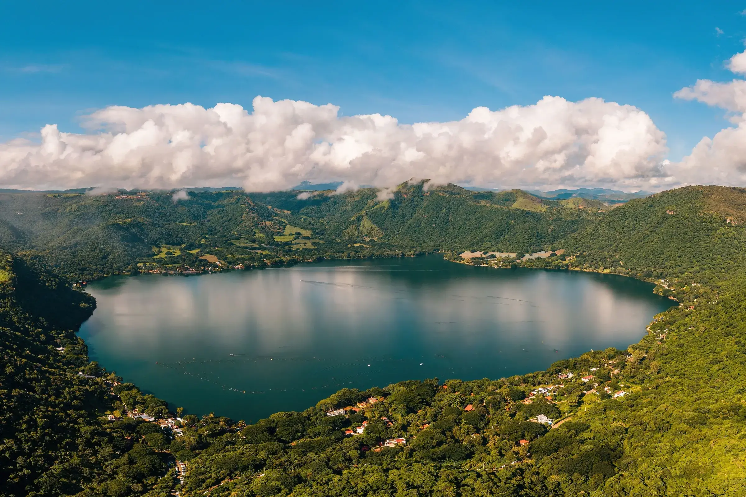 Aerial view of a dark blue volcanic lake surrounded by lush green hills under a bright blue sky with fluffy white clouds. Small houses with orange and white roofs are scattered along the lake's edge, which reflects the sky and clouds.