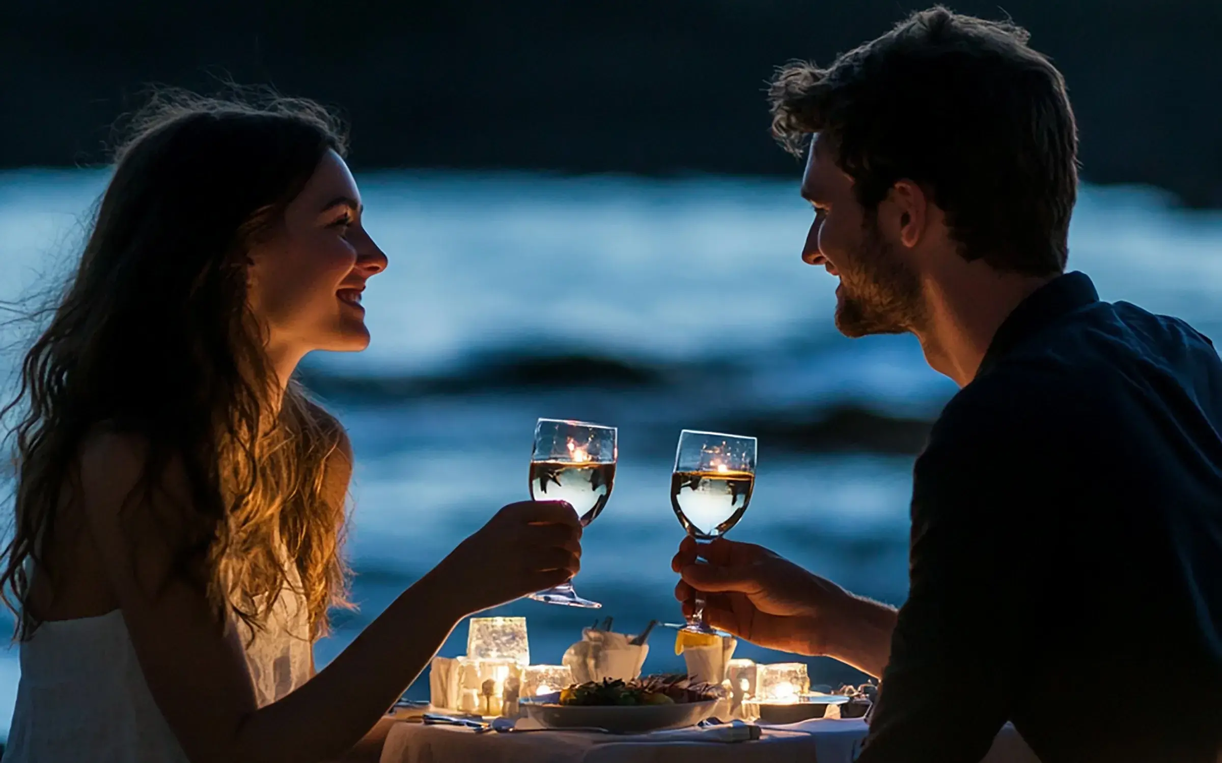 Una pareja sonriente en una cena romántica a la luz de las velas junto al agua por la noche. Están brindando con copas de vino blanco, mirándose con afecto. La luz cálida de las velas ilumina sus rostros y la mesa.