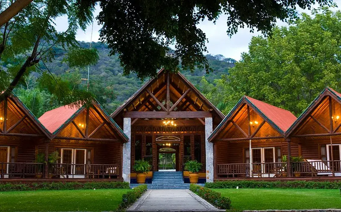 La entrada a un hotel rústico de lujo con varias cabañas de madera y tejados de teja roja. Un camino de piedra atraviesa un césped bien cuidado hacia la entrada principal, que tiene un techo abovedado con vigas de madera. El hotel está enclavado en la base de una exuberante montaña boscosa.