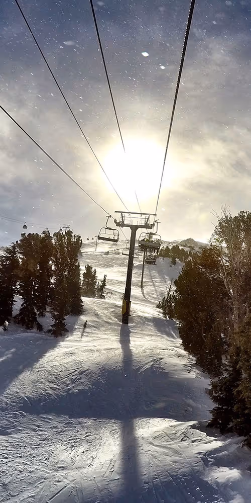 Ski lift chairs ascending a snowy mountain with bright sun and falling snowflakes.