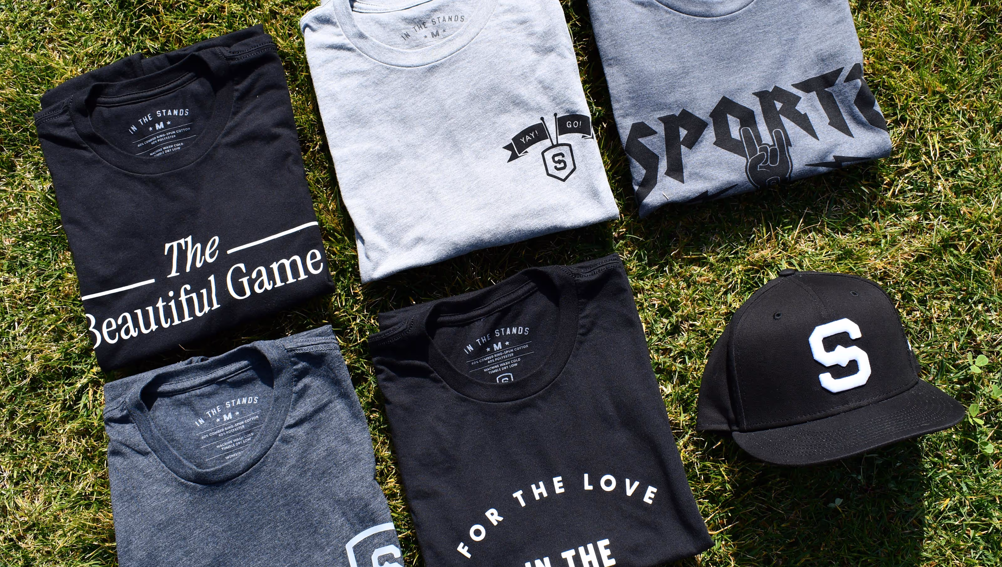 Six folded t-shirts and a black cap with a white letter 'S' on grass, featuring sports-themed prints and logos.