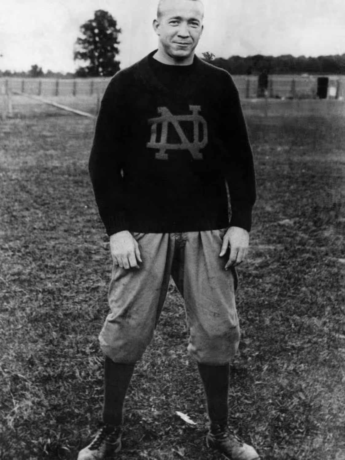 Black and white photo of a man wearing a Notre Dame sweater and old-style football pants standing on a grassy field.