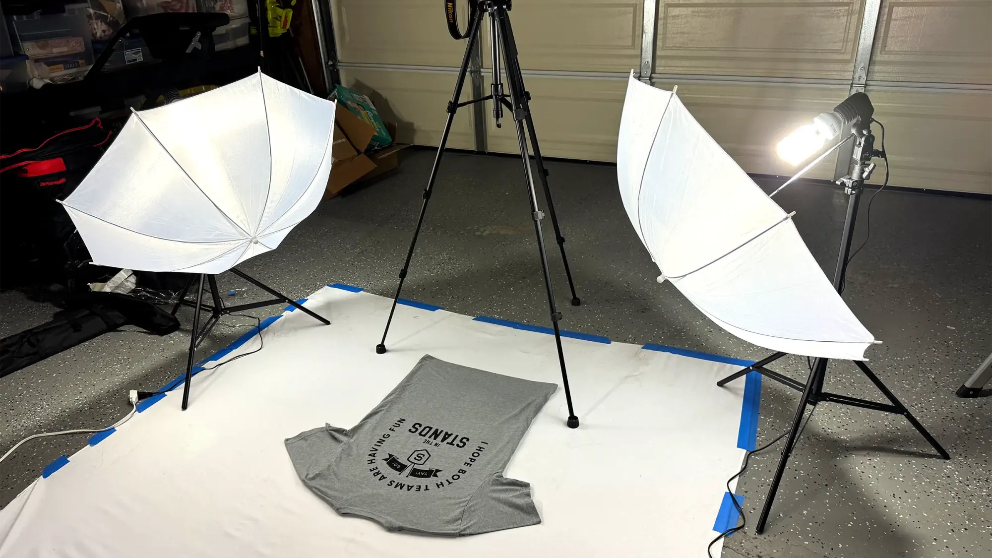 Photography setup with two white shoot-through umbrellas, a tripod, and a gray t-shirt on a white backdrop in a garage.