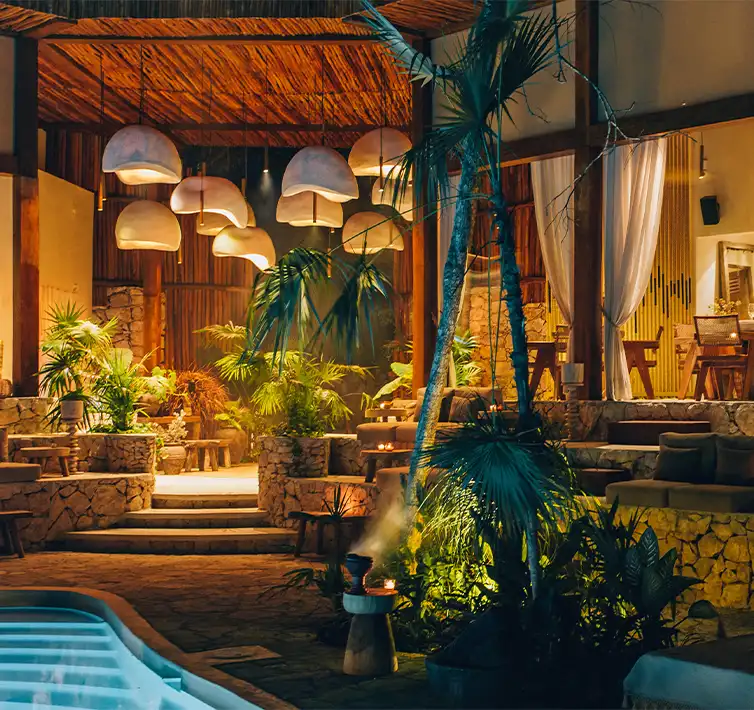 Un acogedor restaurante por la noche, con una iluminación cálida que ilumina las mesas y sillas con el telón de fondo de un edificio con piscina.