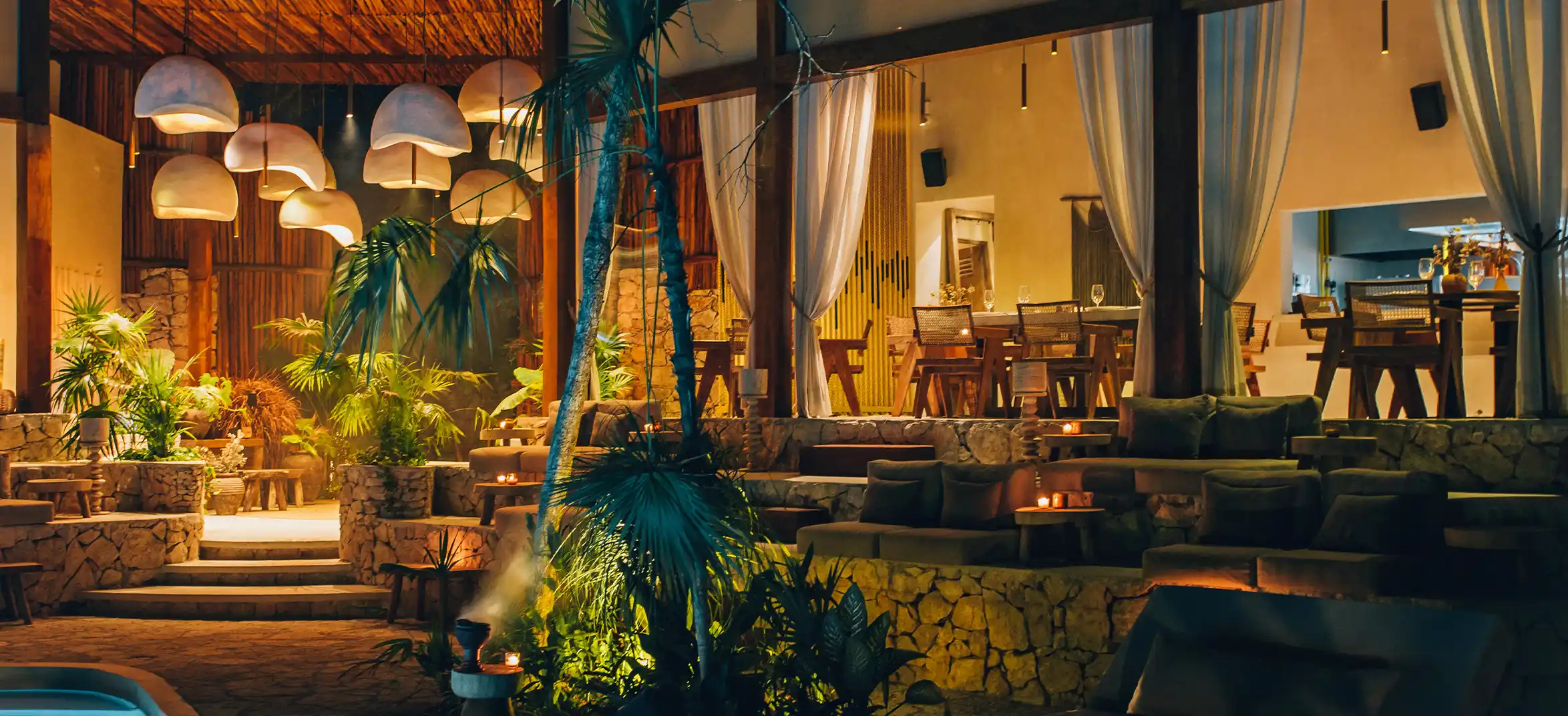 Un acogedor restaurante por la noche, con una iluminación cálida que ilumina las mesas y sillas con el telón de fondo de un edificio con piscina.