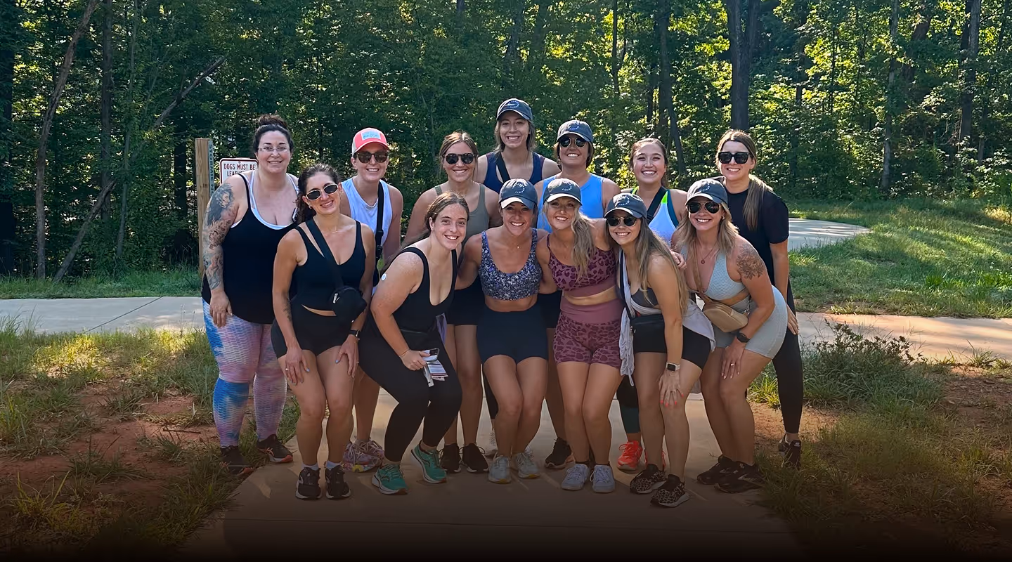 Group of fourteen women dressed in athletic wear posing outdoors on a paved trail with trees in the background.
