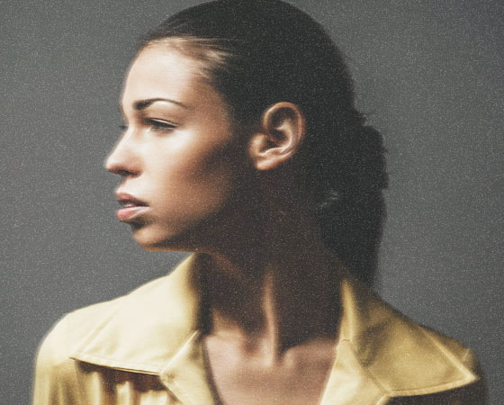  A portrait of a woman in profile, looking to the left. She has dark hair pulled back, and is wearing a yellow jacket with a wide collar. The background is a solid, neutral gray. The lighting highlights her facial features and the sheen of her skin.
