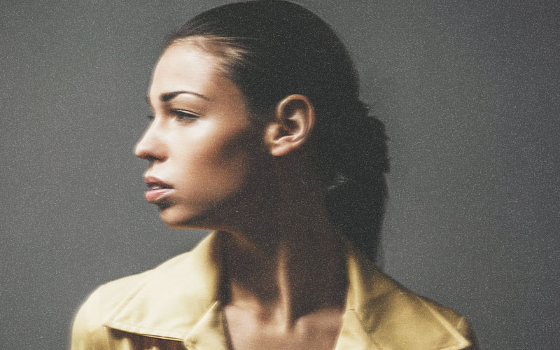  A portrait of a woman in profile, looking to the left. She has dark hair pulled back, and is wearing a yellow jacket with a wide collar. The background is a solid, neutral gray. The lighting highlights her facial features and the sheen of her skin.