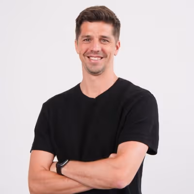 Smiling man with short brown hair wearing a black t-shirt and smartwatch, standing with arms crossed against a white background.