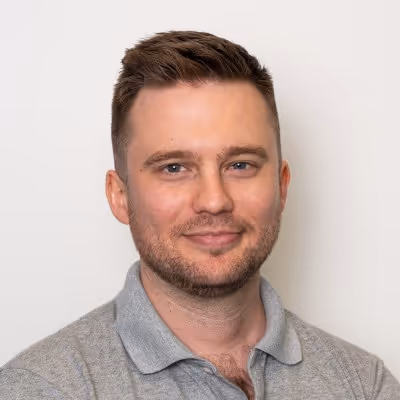 Smiling man with short brown hair and light beard wearing a grey polo shirt against a plain background.