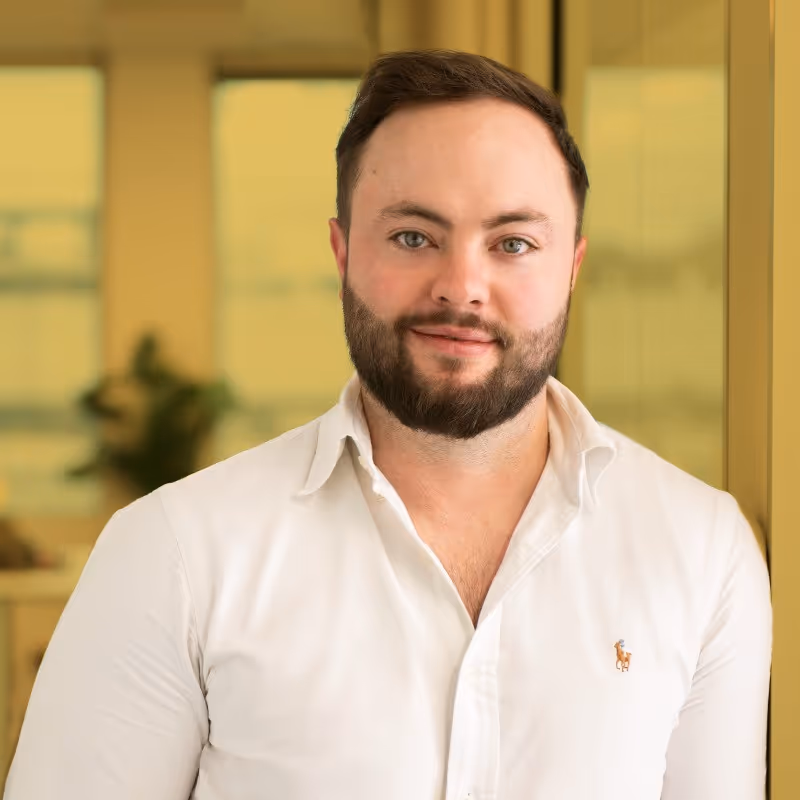 Bearded man wearing a white collared shirt standing indoors with a blurred background.