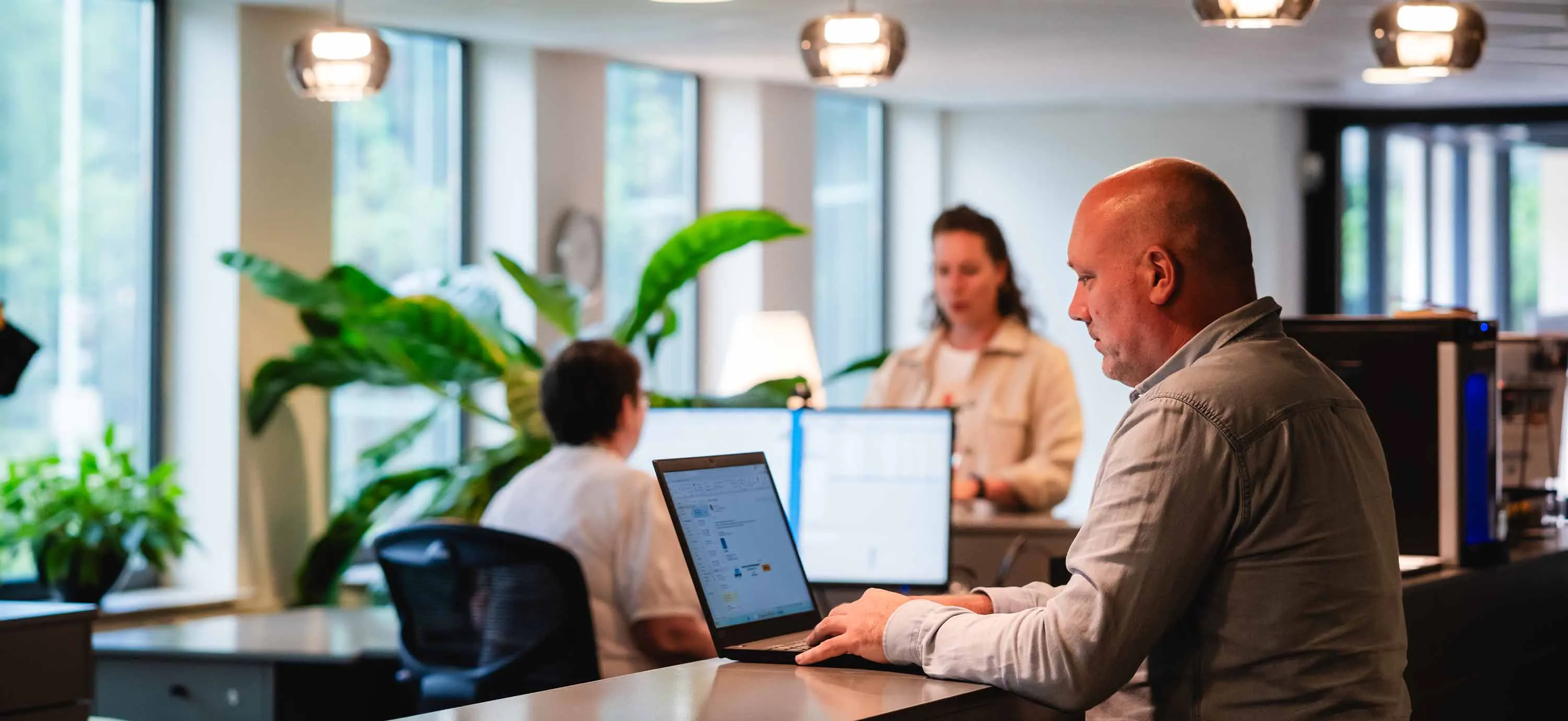 Man werkt op een laptop aan een bureau in een kantoor met twee andere mensen en planten op de achtergrond.
