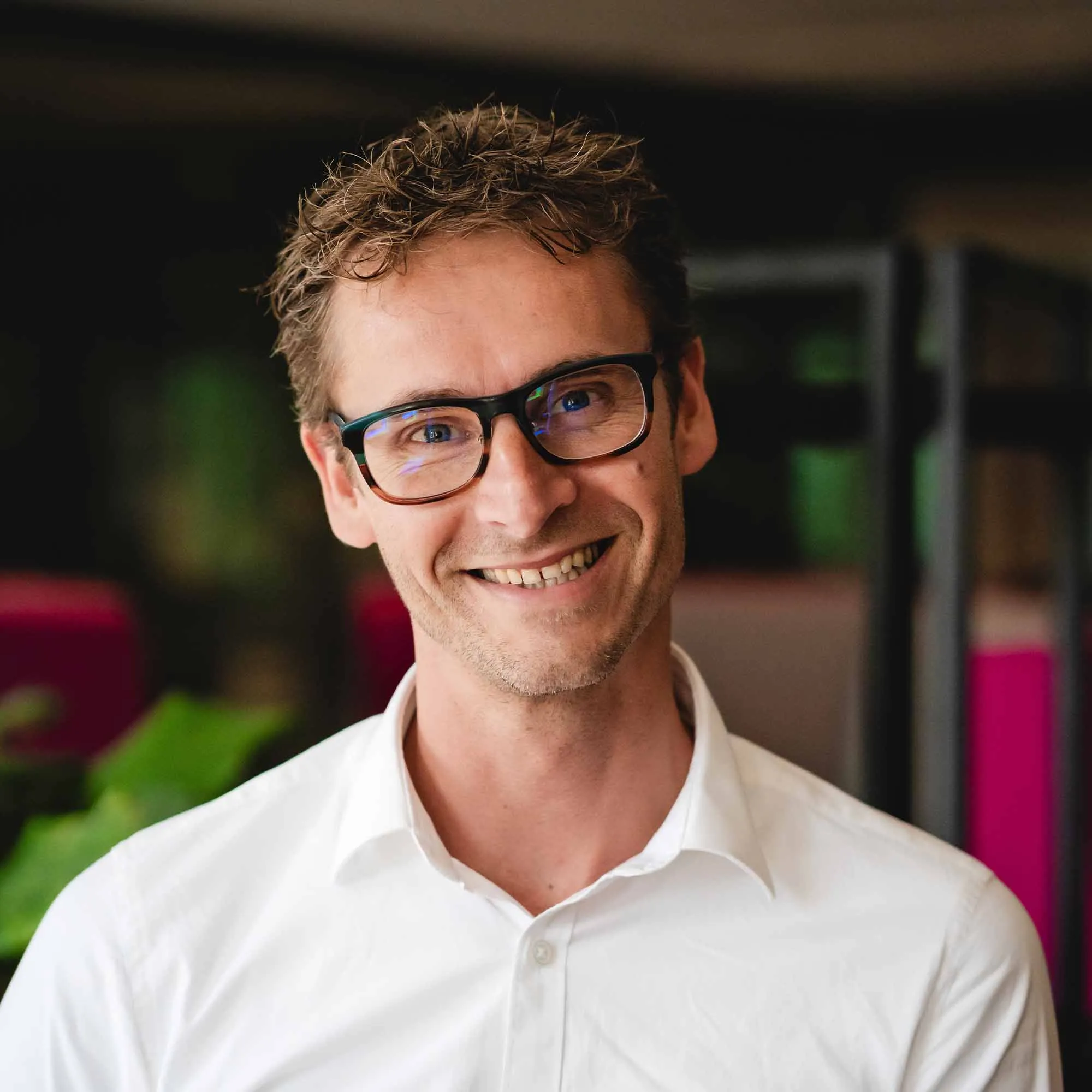 Man met lichtbruine krullende haren en zwarte bril glimlacht, gekleed in een witte overhemd.