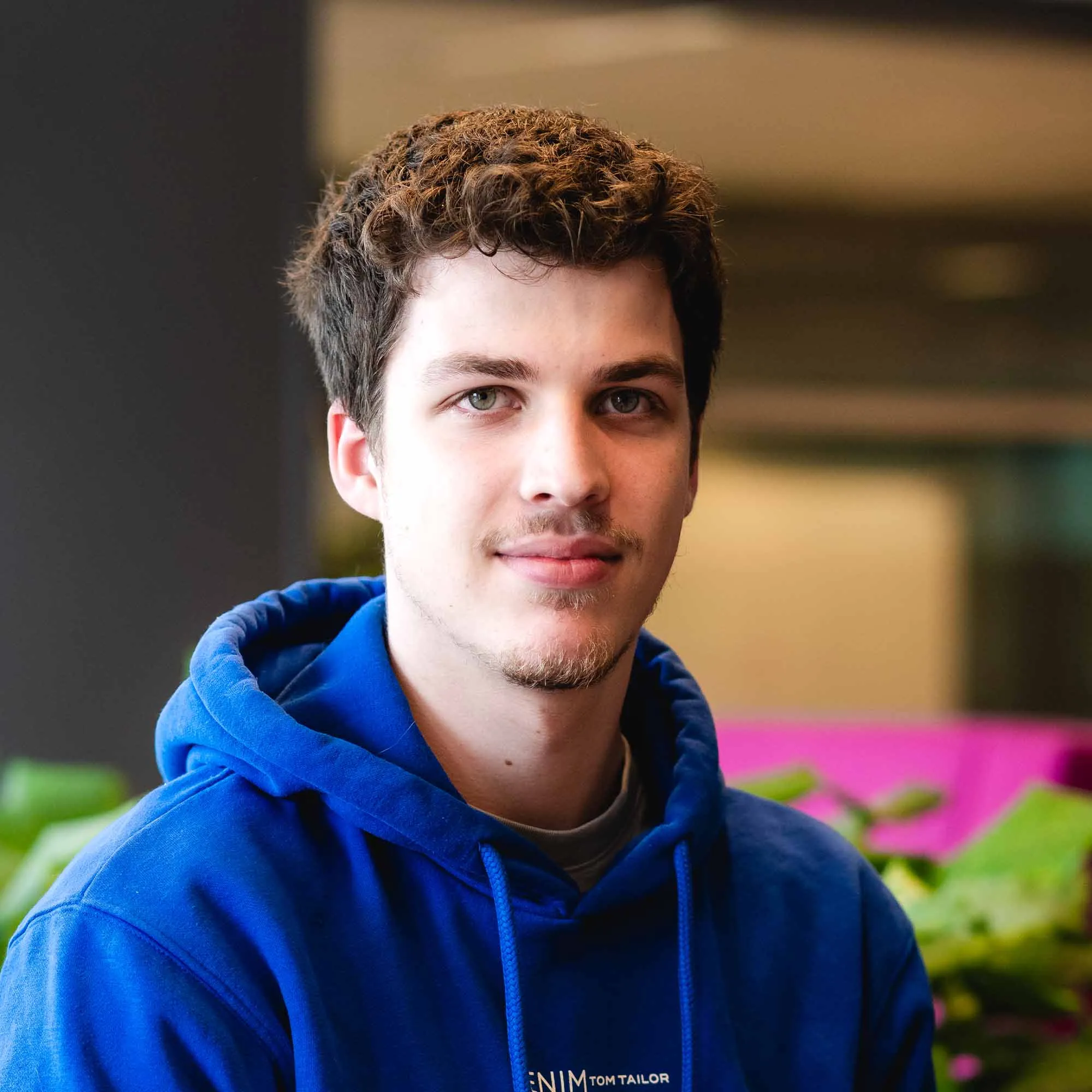 Jonge man met krullend bruin haar en lichte baard draagt een blauwe hoodie en kijkt recht in de camera met een zachte glimlach.