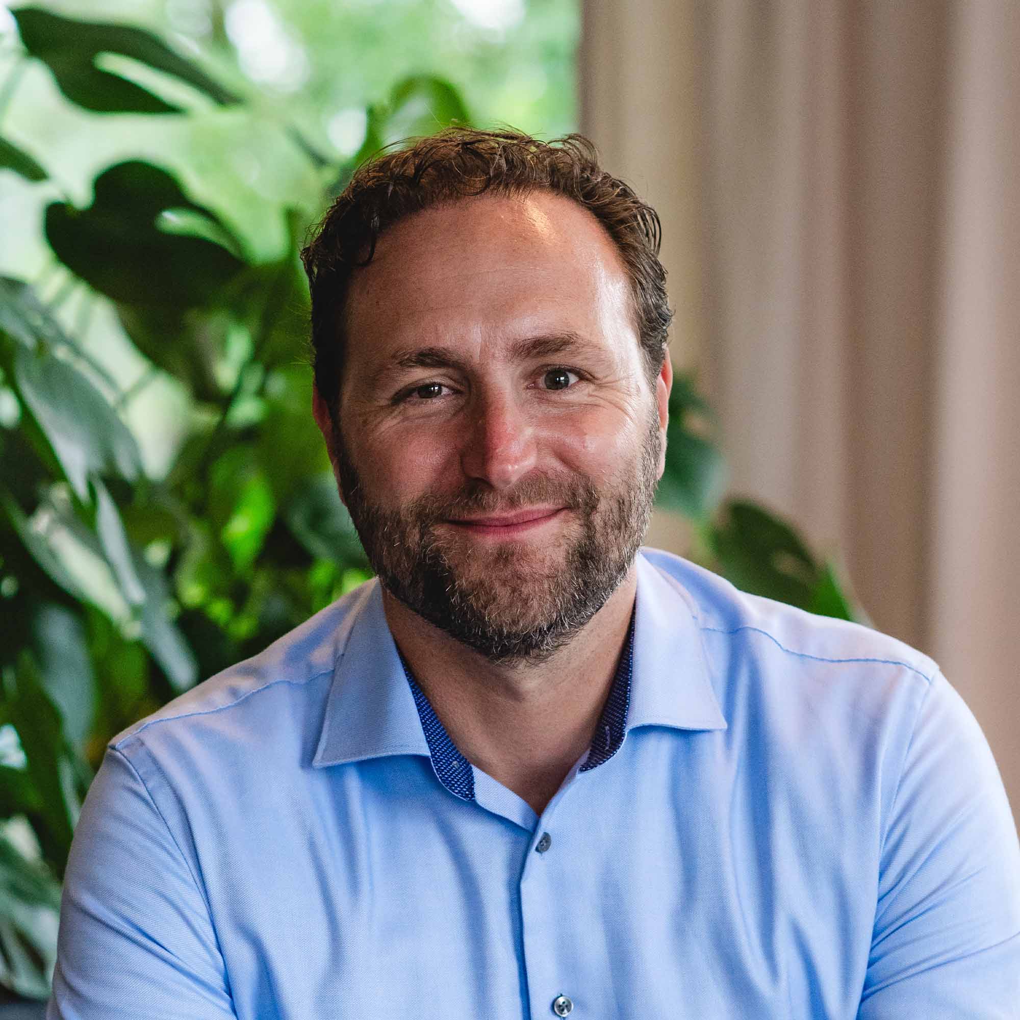 Man met krullend haar en baard, glimlachend in een lichtblauw overhemd met groene planten op de achtergrond.