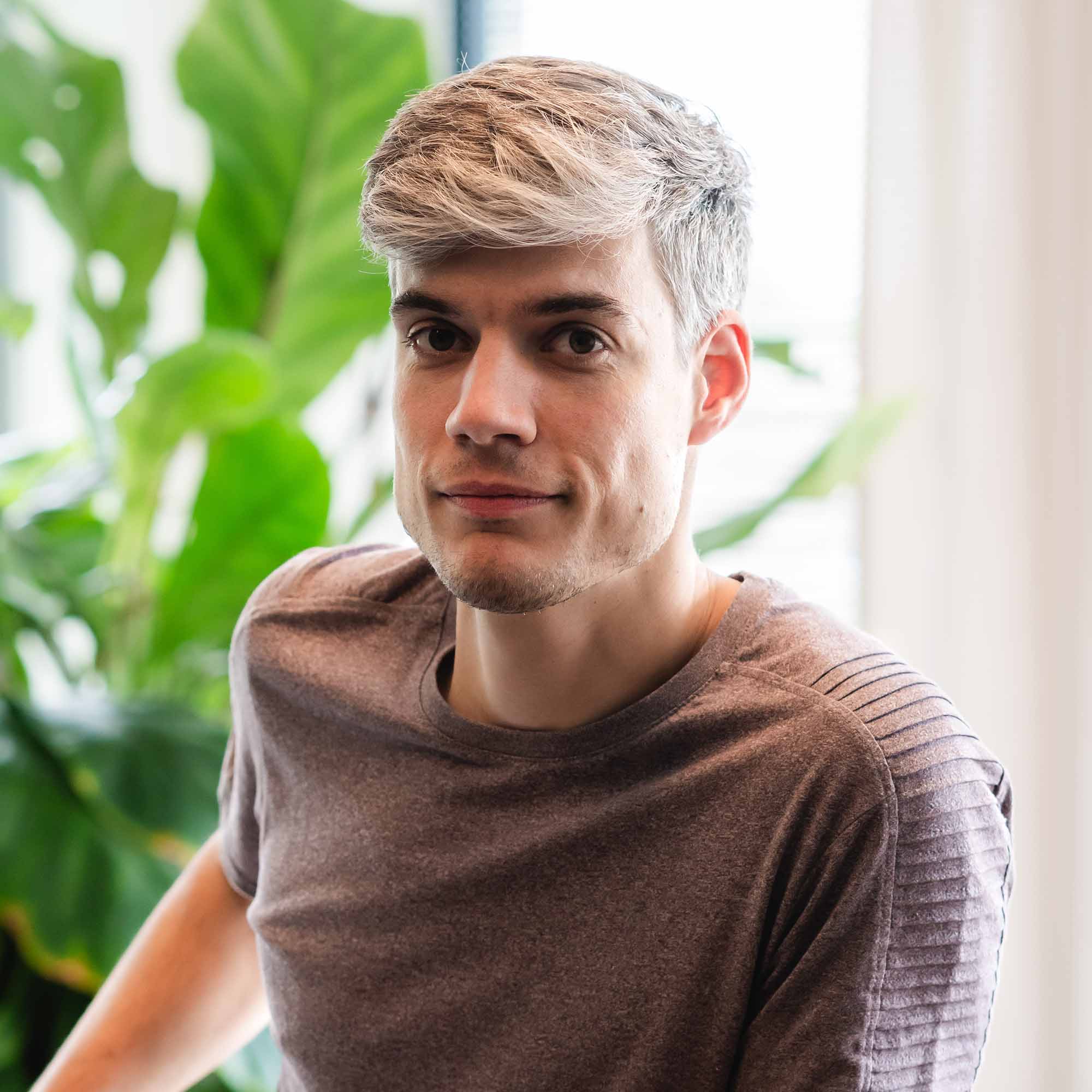 Jonge man met blond haar en bruine T-shirt kijkt recht in de camera, met groene planten op de achtergrond.