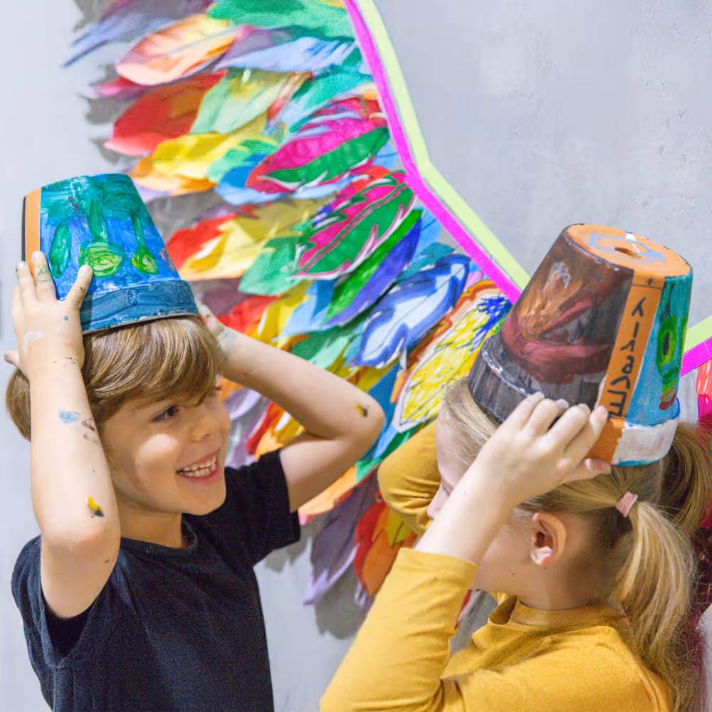 Two children smiling and wearing colorful painted pots on their heads with a colorful feather backdrop.