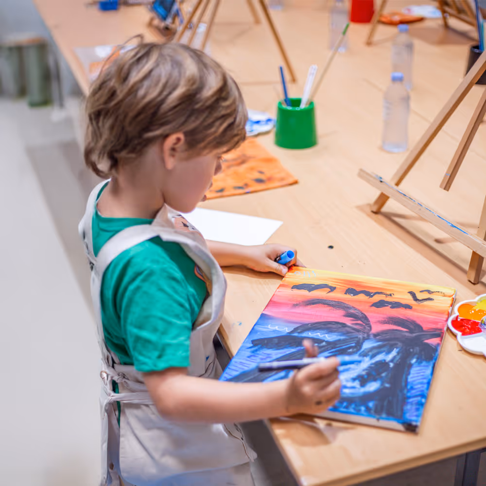 Child painting a colorful sunset with palm trees and flying birds at an art table.