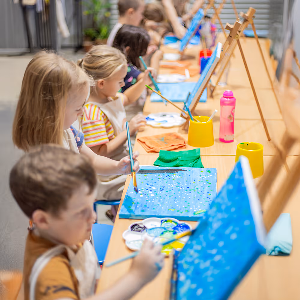 Children sitting at a long table, painting on canvases with brushes and palettes in an art class.