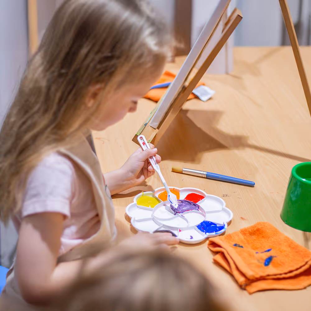 Child mixing purple paint on a palette while sitting at a table with an easel and paintbrush.