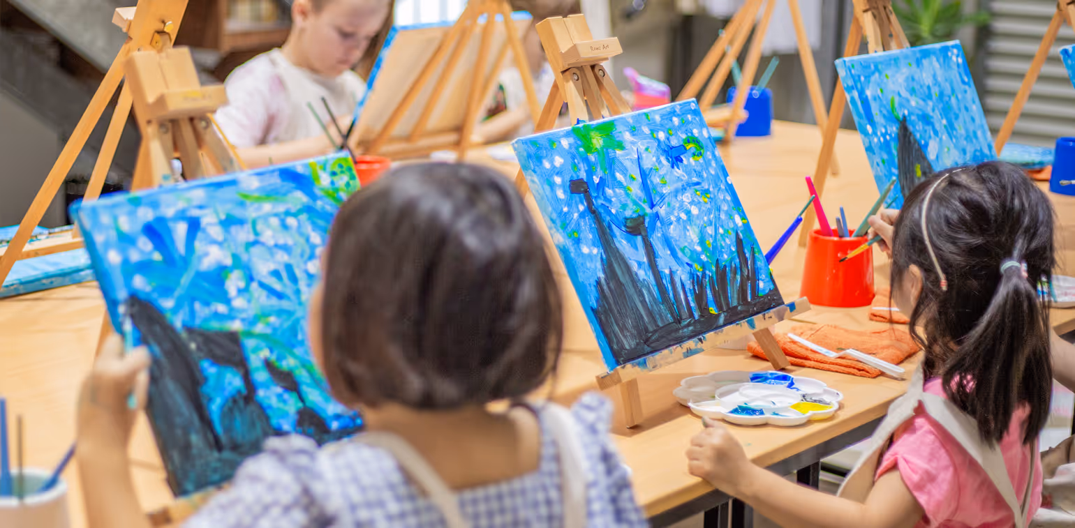 Children seated at tables painting colorful canvases on easels during an art class.