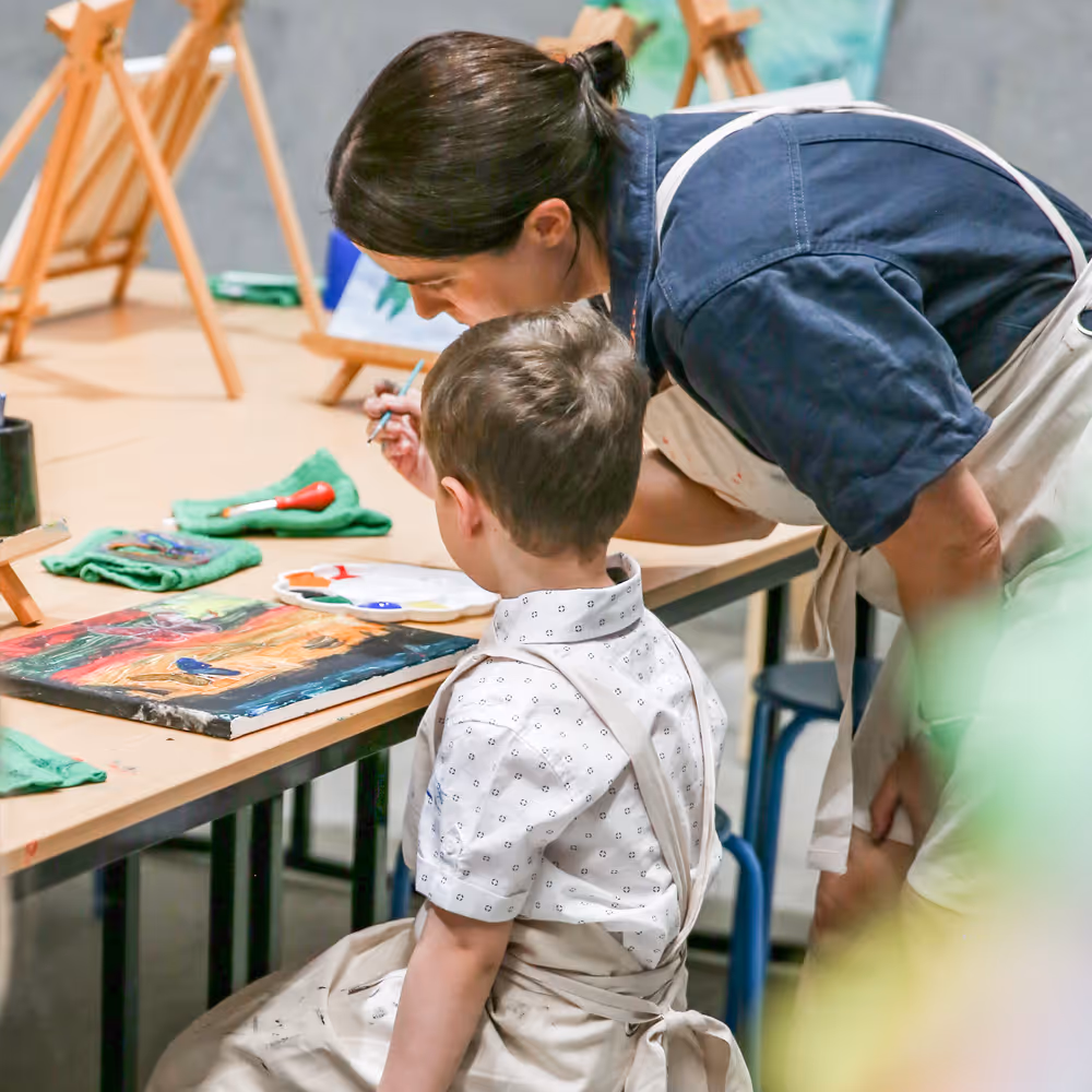 Creative Connection team member helping young boy in apron paint at a table with easels and art supplies.