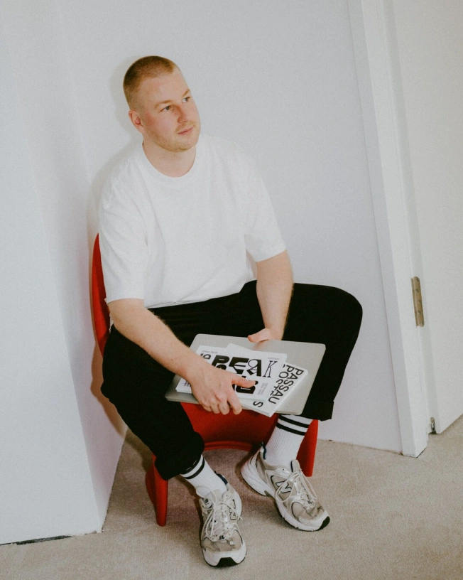 Portrait of Immanuel sitting on a red chair, holding a magazine and a MacBook.