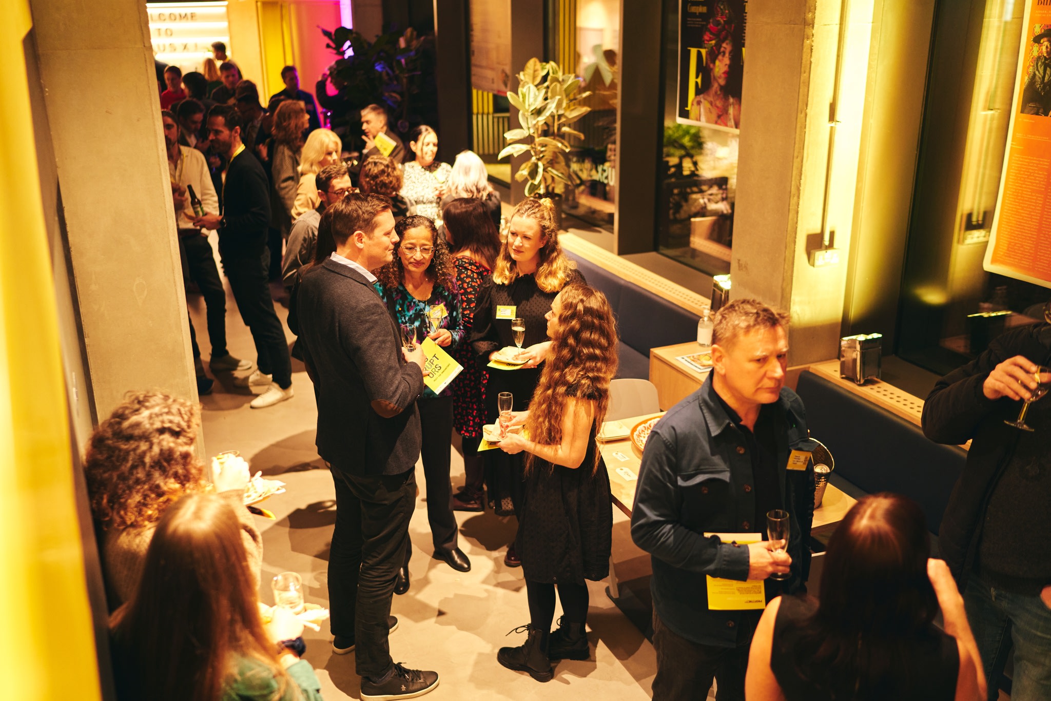 A busy black tie event. The space yellow walls, concrete elements with low lighting. It's focusing on two people chatting, one in a suit and the other in a black dress.
