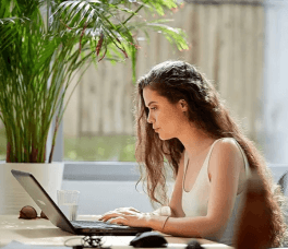 Woman with long hair working over a laptop in a bright space with large plants in the background