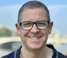 White man with short grey hair and glasses smiling looking at the camera.