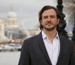 white man with shoulder length hair wearing a suit, in London with St.Paul's in the background