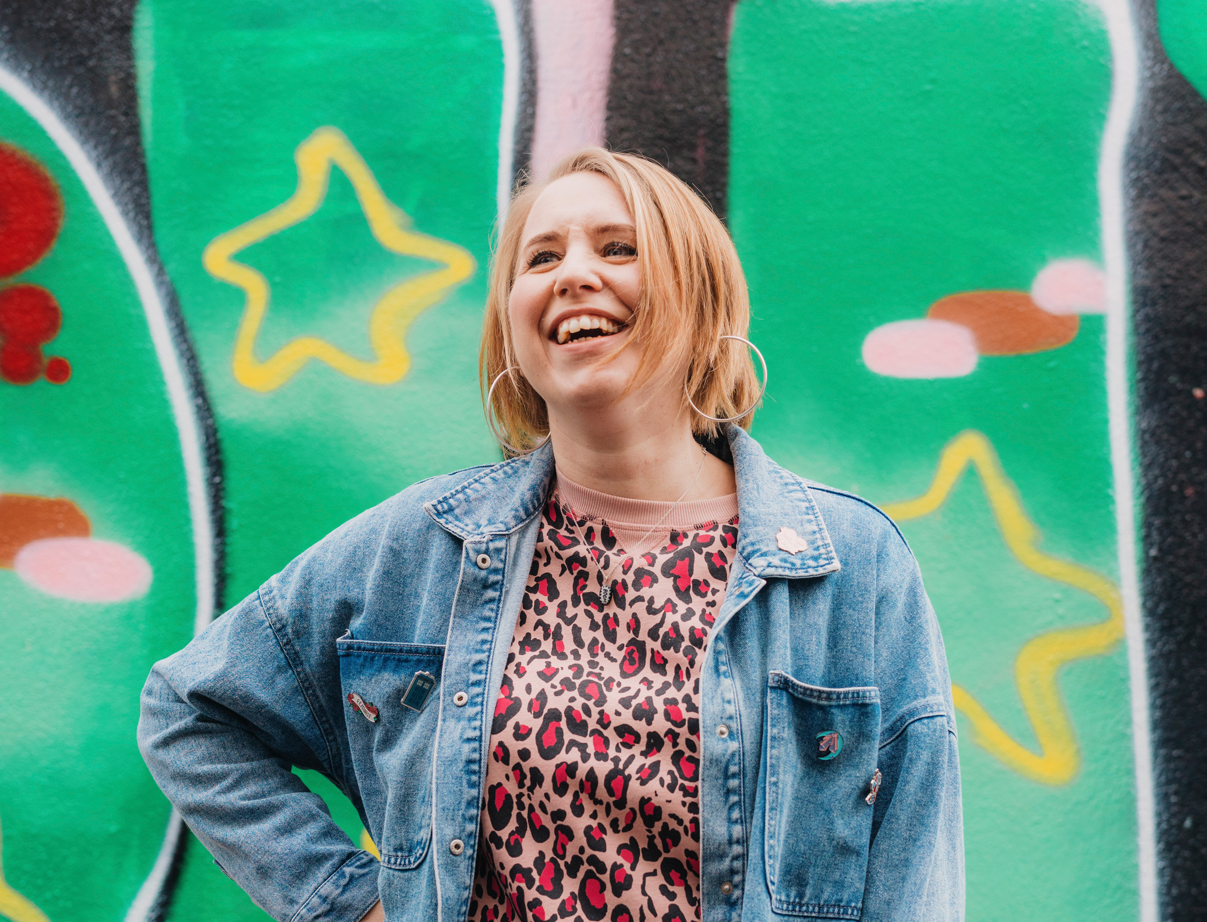 Woman with blonde hair and a denim jacket, looking upwards and laughing. The background is heavily graffitied 
