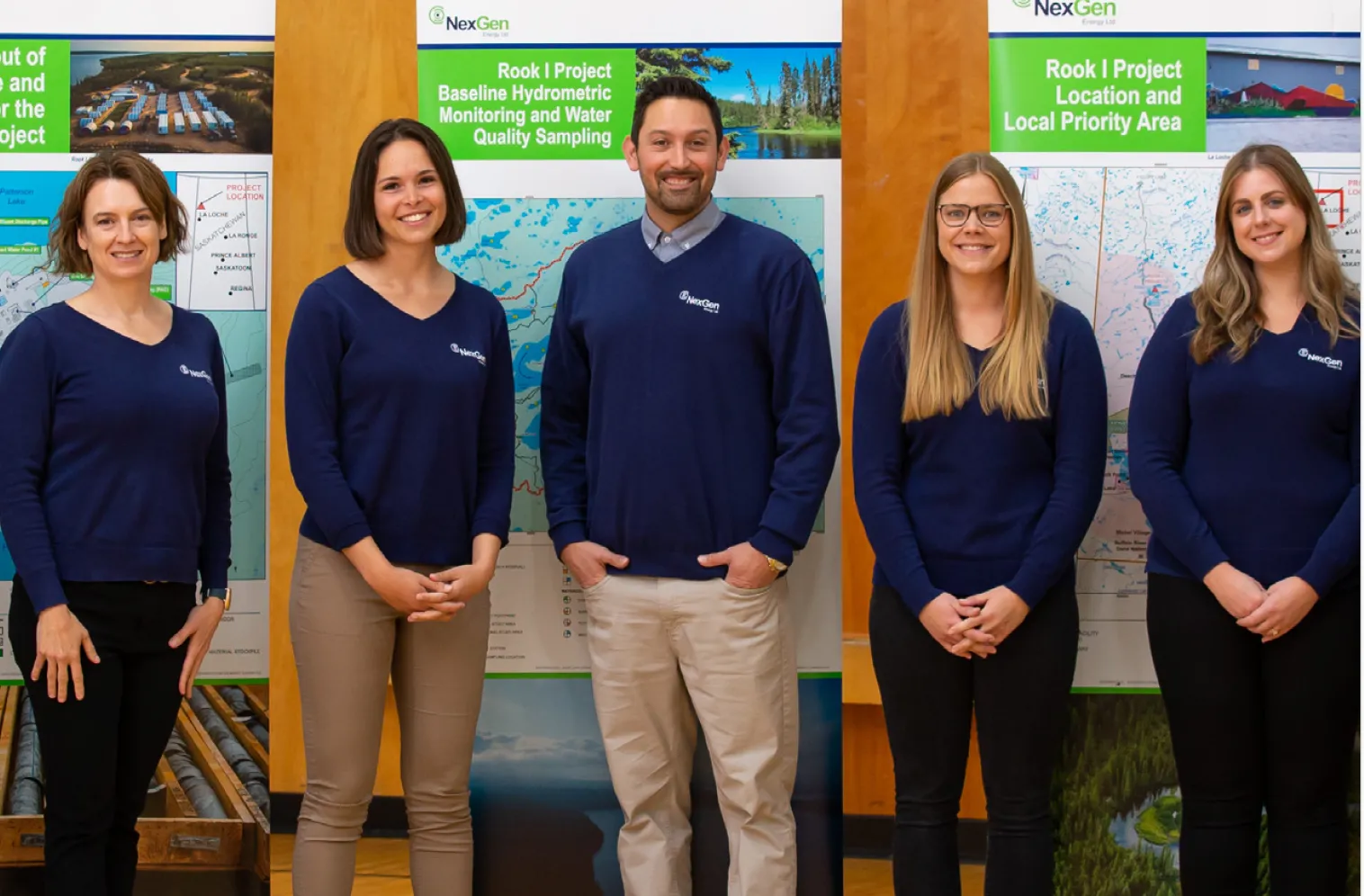 Five people wearing navy NexGen sweaters stand smiling in front of project maps and informational posters.