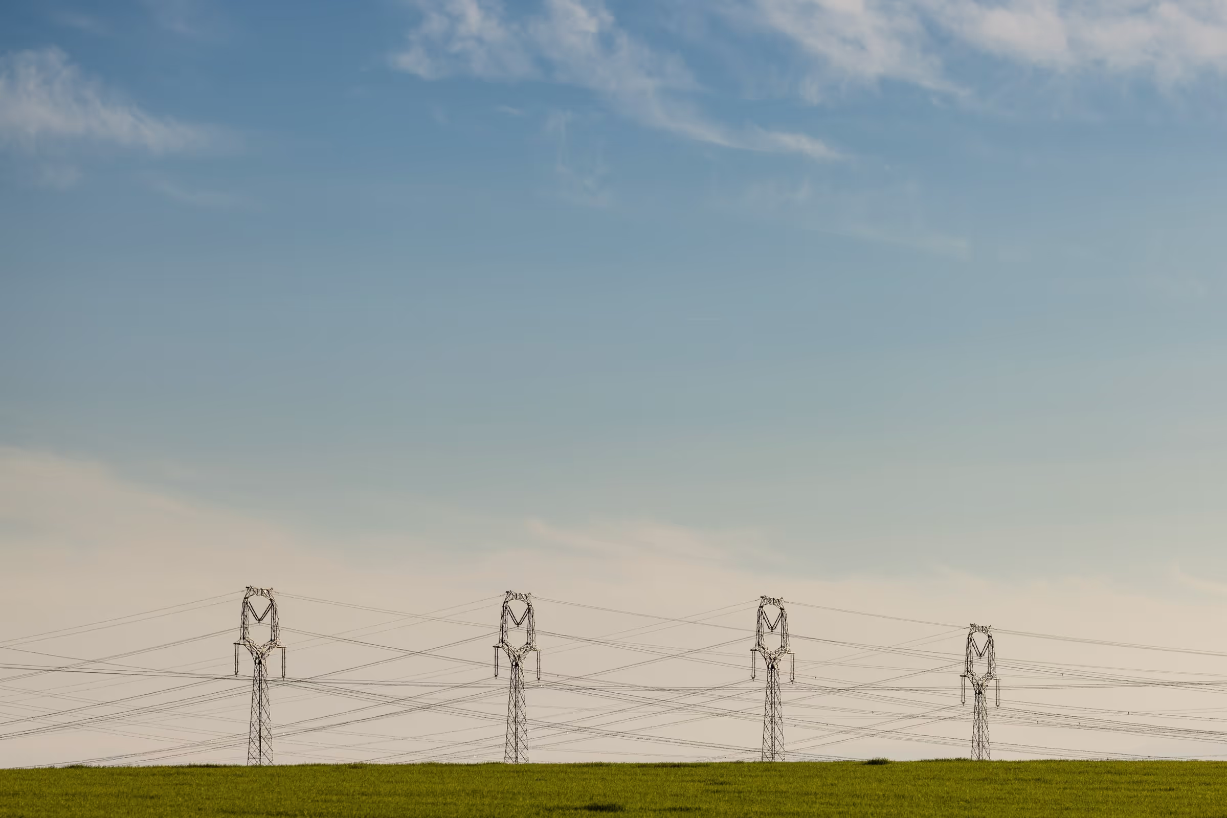 Photo of a clear blue sky with four electricity pylons standing on a grassy field.