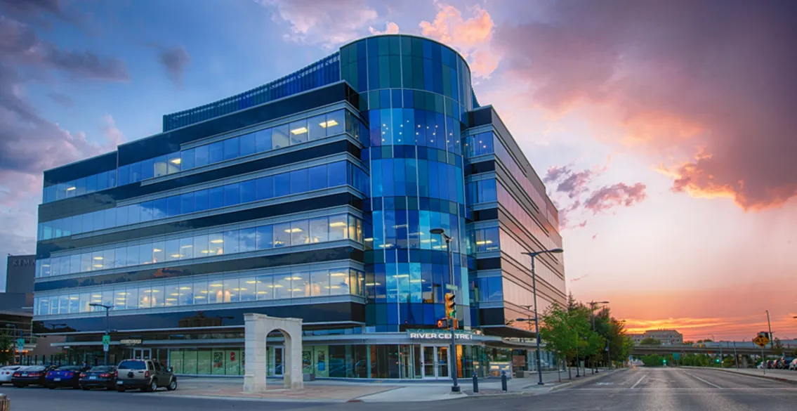 Modern glass office building named River Centre at sunset with street and parked cars.