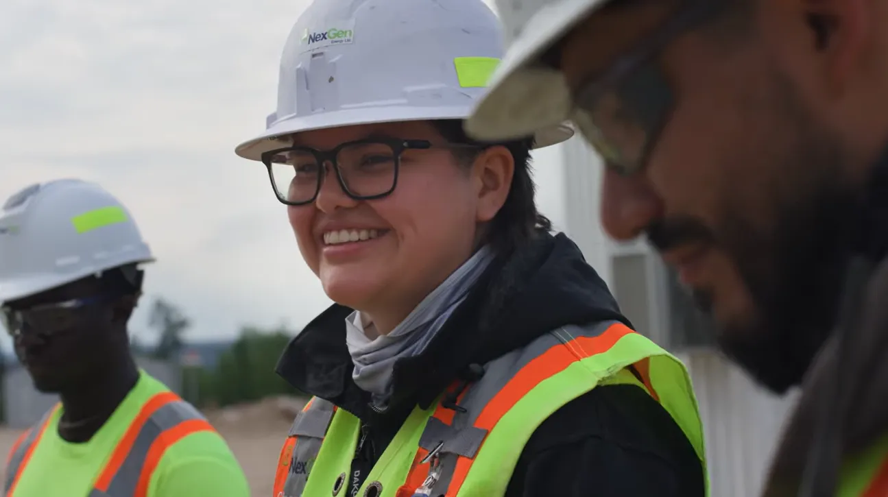 Three construction workers in safety gear, smiling and standing outdoors at a work site.