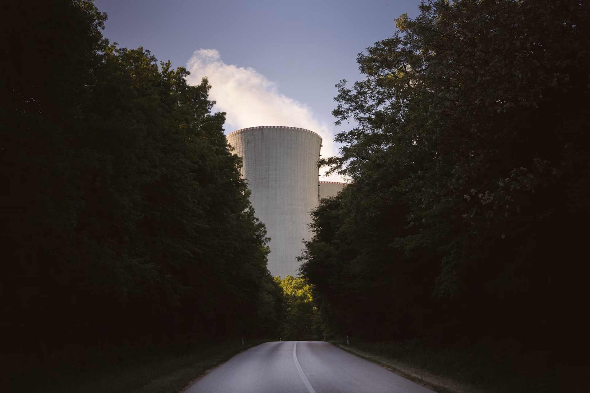 Nuclear power plant cooling tower visible ahead on road flanked by dense green trees under a clear sky.