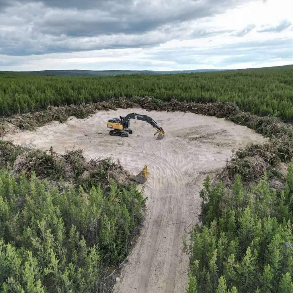 Excavator clearing a circular area in the middle of a dense green forest under a cloudy sky.